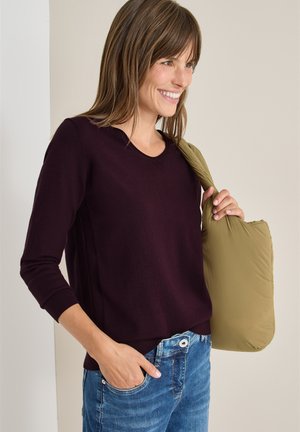 Bordeaux trui met een ronde halslijn, lange mouwen en geribbelde manchetten. Gepaard met blauwe jeans; houdt een licht olijfgroene puffer tas vast.