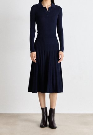 Femme portant une robe en maille côtelée bleu marine à manches longues avec une jupe plissée et des bottines noires, debout contre un mur blanc.