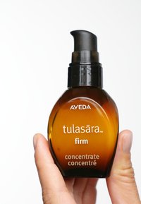 Bouteille en verre ambre avec un bouchon à pompe noir. L'étiquette avant indique "Aveda tulasāra concentré raffermissant." Le texte est blanc et clairement lisible.
