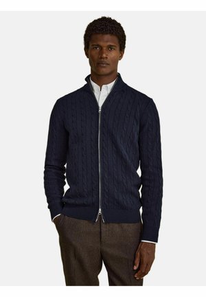 Mand iført en navy blå kabelstrikket sweater med lynlås over en hvid skjorte og brune bukser, stående med hænder delvist i lommerne.