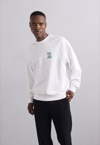Weißer Pullover mit Rundhalsausschnitt und langen Ärmeln; mit grünem Logo auf der linken Brust; kombiniert mit schwarzen Hosen. Glatte Textur.