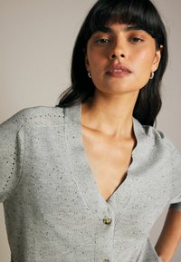 Grå strikket cardigan med korte ærmer, med en dyb V-hals, to knapper og subtil spraglet tekstur i hele stoffet.