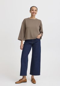 Felpa marrone over size con maniche ampie, abbinata a jeans a gamba larga blu scuro e mocassini marroni. Tessuto liscio, design semplice.