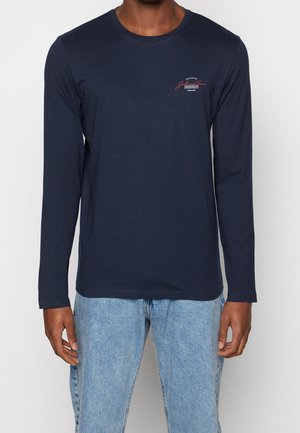 Homme portant une chemise à manches longues bleu marine avec un petit logo texte rouge et blanc sur le côté gauche de la poitrine, associée à un jean bleu clair.