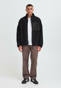 Man standing wearing a black fleece jacket over a white shirt, brown pants, and black sneakers with white stripes against a plain background.