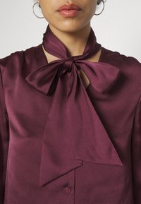 Femme portant une blouse en satin violet foncé avec un grand nœud noué au cou et des boucles d'oreilles argentées en forme de créoles, lèvres visibles mais pas les yeux.