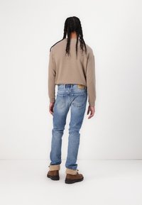 Lichtbruine langemouwshirt, blauwe spijkerbroek en bruine laarzen met bontvoering. Opvallende stiksels en label op de spijkerbroek. Achteraanzicht.