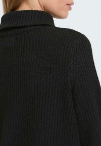 Pulover negru din tricot cu o guler înalt, pliat și o suprafață texturată. Designul prezintă linii verticale și o croială lejeră.
