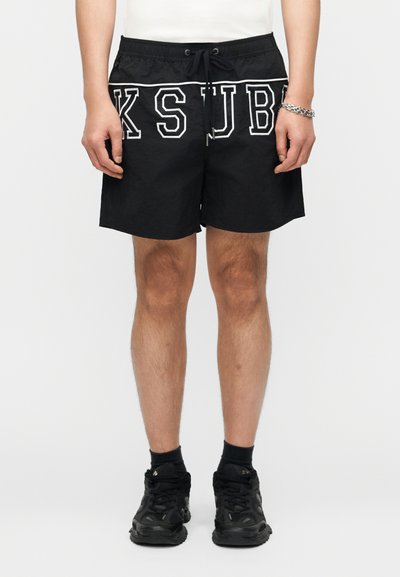 Homme portant un short noir avec les lettres KSJB en contour blanc, un T-shirt blanc, des chaussettes noires, des baskets noires et un bracelet chaîne en argent.