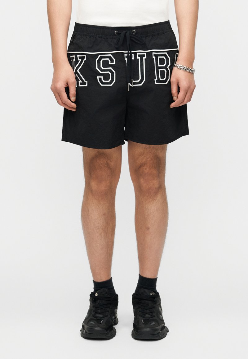 Vyras dėvi juodus šortus su baltais kontūruotais raidėmis KSJB, baltą marškinėlį, juodas kojines, juodus sportinius batelius ir sidabrinį grandinėlės apyrankę.