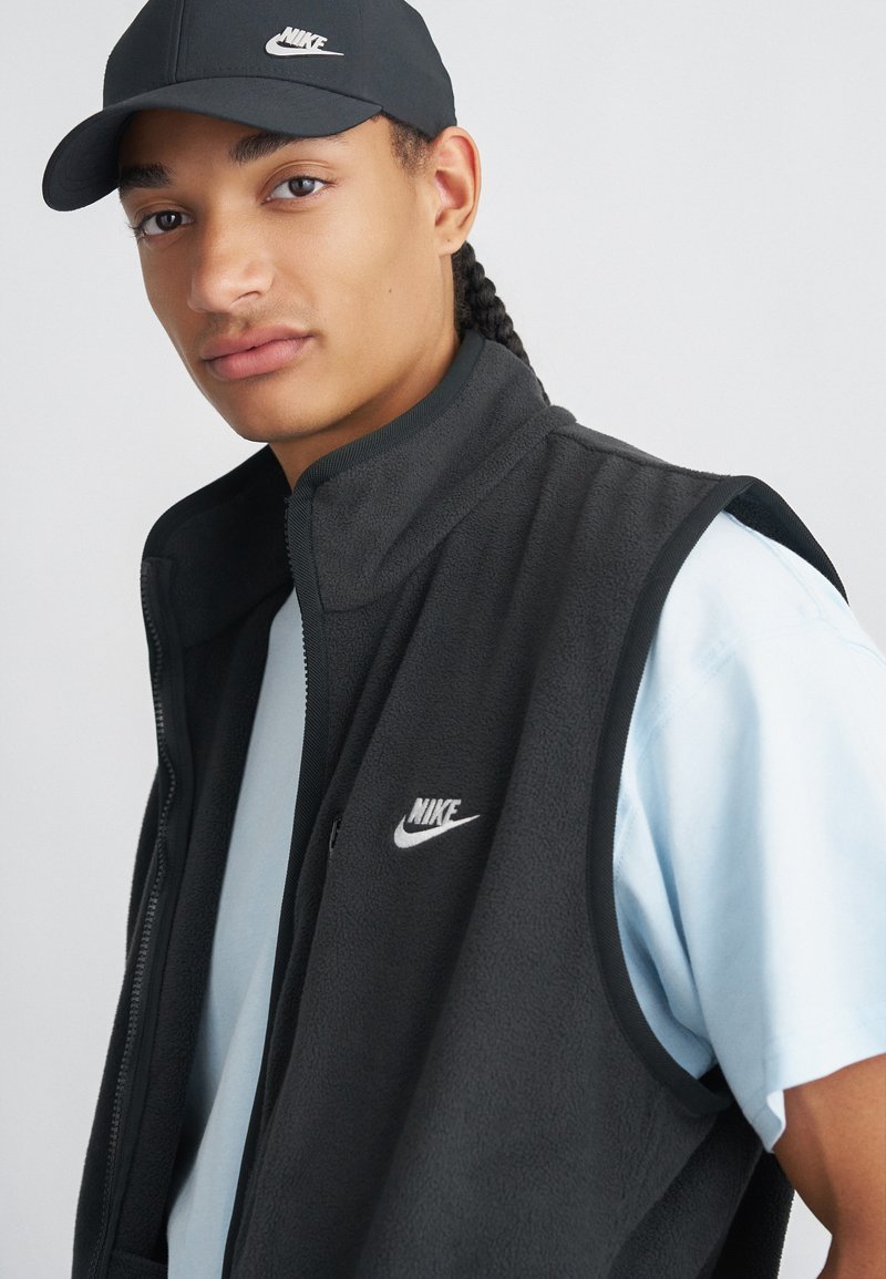 Vest din fleece negru cu guler înalt, având un logo alb Nike. Cămașă albastră deschisă dedesubt, asortată cu o șapcă neagră.