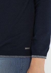 Námořnický modrý svetr s rukávy a lemem se stříbrným lemováním, částečně zakrývající světlé modré džíny, s malou kovovou značkou blízko lemu.