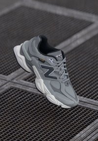 Graue und weiße New Balance Sneaker mit "9060"-Aufschrift und Cordura-Stoffetikett, platziert auf einer Metallgitteroberfläche.