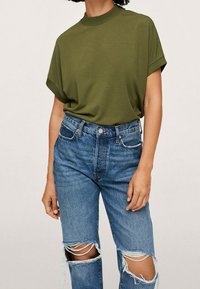 Oliwkowa dziergana bluzka z krótkim rękawem i wysokim kołnierzem, w połączeniu z przetartymi niebieskimi dżinsami o klasycznym kroju z postrzępionymi kolanami.