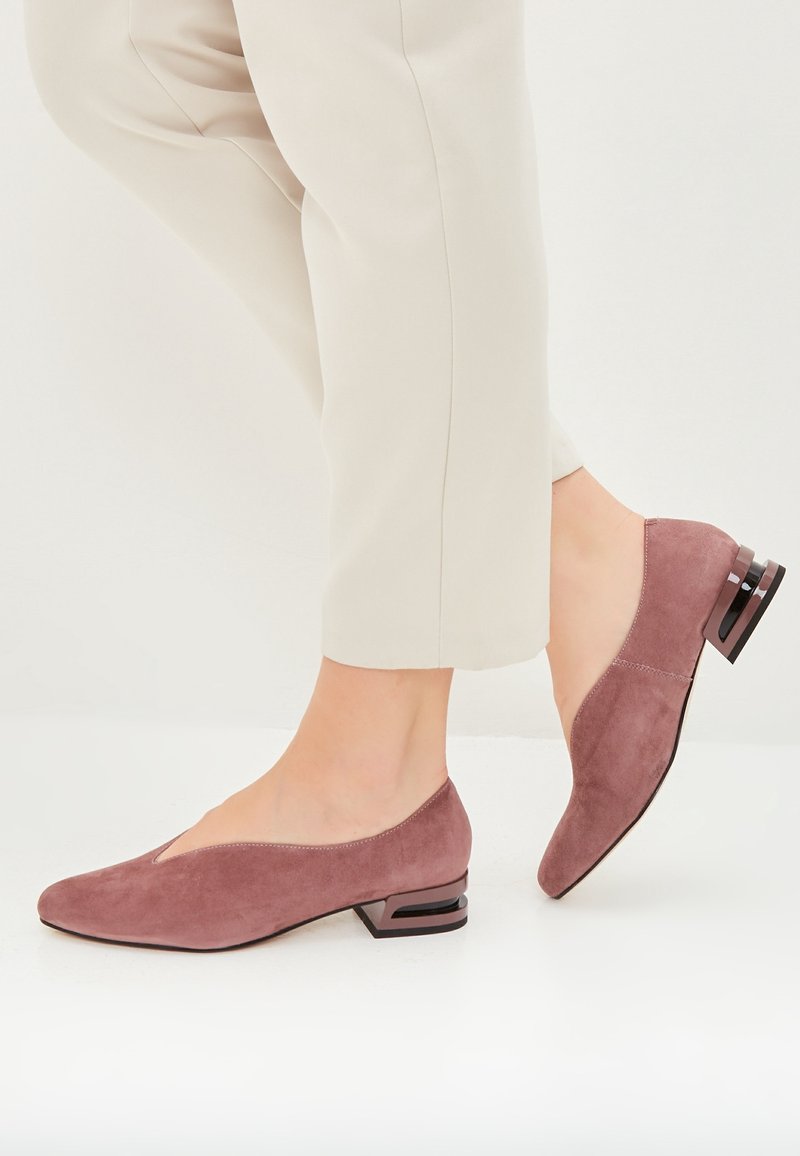 Person iført beige cropped bukser og mauve suede lavhælede sko med en unik udskæring på hælen, stående på et hvidt gulv.