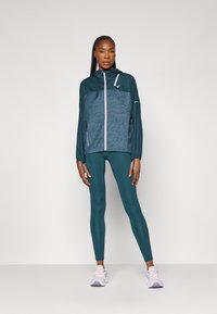 Tyrkis hættetrøje med tekstureret mønster, pink lynlås og letvægtsdesign, parret med matchende tyrkise leggings og lyse sneakers.