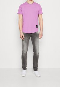 Osoba v světle fialovém tričku Calvin Klein, vybledlých šedých slim-fit džínách, bílých teniskách a stříbrném řetízku, stojící proti bílé zdi.