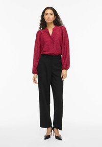 Mørkerød teksturert bluse med knapper foran, kombinert med høytlivede svarte bukser. Modellen har på seg svarte spisse hæler.