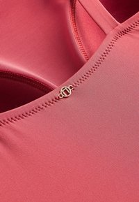 Gros plan sur un tissu rose avec des coutures détaillées, un intérieur en maille et un petit logo circulaire en métal de la marque près de la couture.