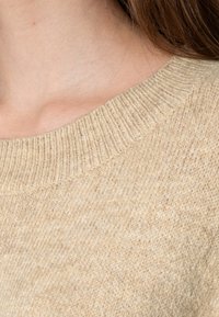 Beige strikket genser med ribbestrikket rund hals og fin tekstur, som viser en nærbilde av stoffet og kragedetaljene.