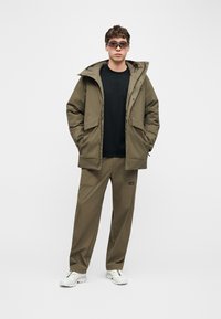 Άνδρας που φοράει ελιάς-πράσινο μπουφάν με κουκούλα και παντελόνι, μαύρο πουκάμισο, λευκά αθλητικά παπούτσια και φιμέ γυαλιά ηλίου, στέκεται μπροστά σε απλό φόντο.