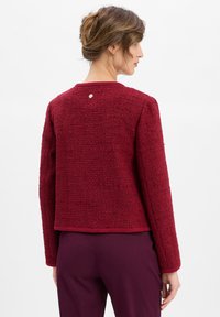 Giacca rossa a trama con maniche lunghe, scollatura rotonda e dettaglio di bottone visibile sul retro; rifinita con un bordo solido all'orlo.