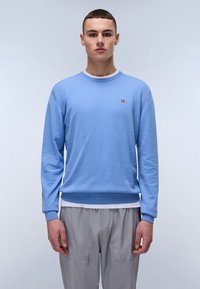 Suéter azul claro hecho de tela suave, con cuello redondo y puños de canalé. Pequeño logo en el lado izquierdo del pecho. Usado sobre pantalones grises.