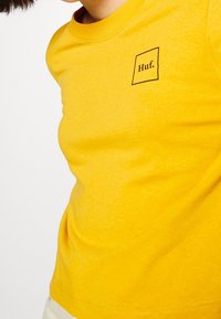 Gul bomullströja med en fyrkantig logotyp där "Huf." står tryckt i svart. Kort ärm och rund halsringning med en mjuk yta.