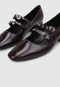 Zapatos de charol marrón oscuro con punteras afiladas, que cuentan con dos correas con herrajes de plata y recortes circulares en los laterales.