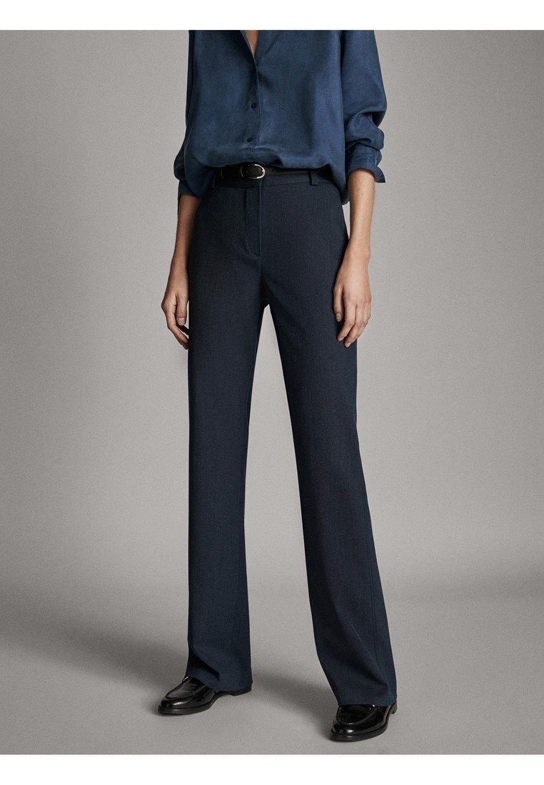 Personne portant un pantalon large bleu foncé, une ceinture noire, une chemise bleu foncé à manches longues et des mocassins noirs, debout devant un fond uni.
