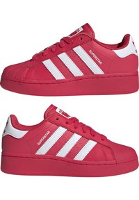 Röda Adidas Superstar-sneakers med vita ränder, snäppe-design och "Superstar"-text på sidan, visade från både vänster och höger vinkel.