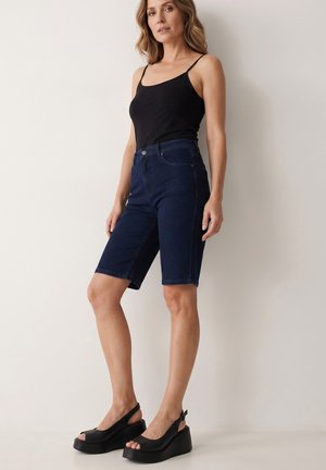 Kvinde stående iført sort ærmeløs top, mørkeblå knælange denimshorts og sorte platform-sandaler mod en ensfarvet baggrund.