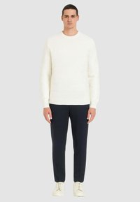 Maglione lavorato a maglia color crema con motivo geometrico testurizzato, abbinato a pantaloni blu scuro navy e sneakers bianche, presentato su uno sfondo neutro.