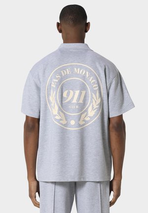 Mann trägt hellgraues Kurzarmhemd und passende Hose mit dem Logo "Pas de Monaco 911 Club" in Beige auf der Rückseite des Hemdes.