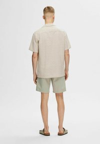 Korte mouwen shirt in licht beige met een herhalend geometrisch patroon, gecombineerd met lichtgroene shorts en bruine sandalen.
