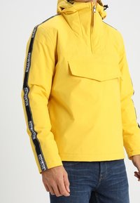 Giacca impermeabile gialla con zip frontale a mezza lunghezza, tasca anteriore e strisce nere sui lati con testo del logo. Cappuccio e polsini regolabili.