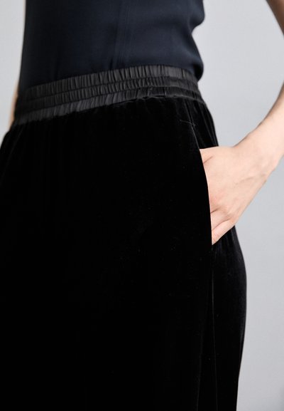 Fustă din catifea neagră cu talie elastică. Are o buzunar pe lateral și o textură netedă, subliniind o siluetă elegantă și fluidă.