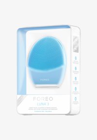 Dispositif de nettoyage facial en silicone bleu avec surface texturée, forme ovale et base transparente, emballé dans une boîte claire avec les détails du produit.
