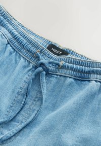 Heledad sinine teksapükste lühikesed püksid, millel on elastne vöökoht, nöördetail ja must "NEXT" silt vöökoha lähedal. Sile tekstuur.