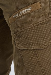 Bruine broek met patroon en een gestructureerde afwerking, voorzien van een zichtbaar logo op de zak met de tekst "PME LEGEND AMERICAN CLASSIC".