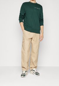 Grön bomullströja med rund hals och GANT-logotyp, tillsammans med beige cargobyxor med sidofickor och gröna och vita sneakers.