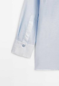 Chemise bleu clair à manches longues avec un poignet à un seul bouton, montrant une partie de la manche et la couture latérale sur un fond uni.