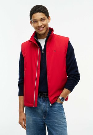 Ung mand smiler iført en rød ærmeløs jakke over en marineblå sweater og blå jeans, stående mod en hvid baggrund.