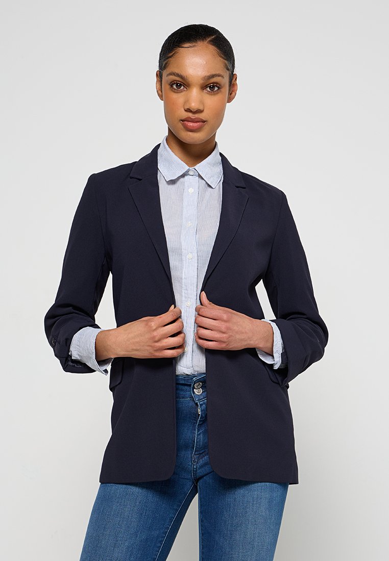 Blazer blu navy con chiusura a un bottone, colletto a revers e due tasche, indossato sopra una camicia a righe chiara con bottoni e jeans in denim.