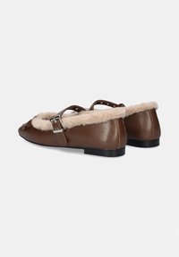 Mocasines de piel marrón con un suave forro de piel de oveja beige, correas al tobillo con herrajes plateados y un bajo tacón negro. Superficie lisa y punta redondeada.