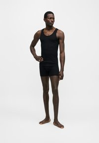 Άνδρας μοντέλο όρθιος ξυπόλυτος, φορώντας μαύρο αμάνικο τοπ και μαύρα εφαρμοστά σορτς σε απλό λευκό φόντο.