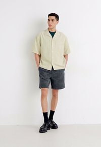 Limoengroene gestructureerde kortebloes, met een voorzak, gecombineerd met donkergrijze shorts en zwarte schoenen, gedragen met zwarte sokken.