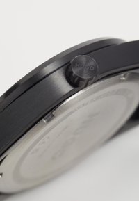 Melna nerūsējošā tērauda pulkstenis ar teksturētu rāmīti un izteiksmīgu kronīti, kas marķēts ar "HUGO". Iespējo minimālistisku ciparnīcu ar sudraba akcentiem.