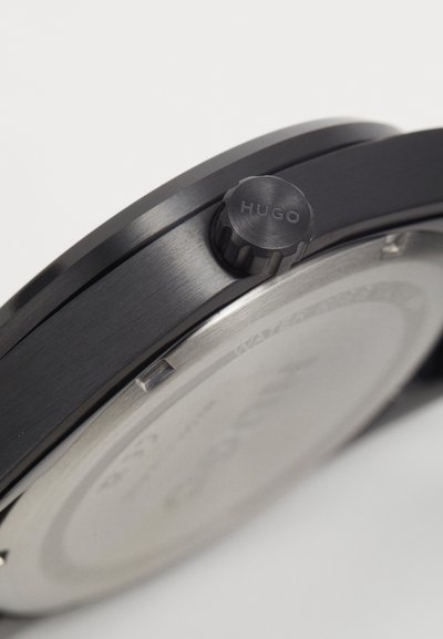 Montre en acier inoxydable noir avec une lunette texturée et une couronne proéminente étiquetée "HUGO". Présente un cadran minimaliste avec des accents en argent.