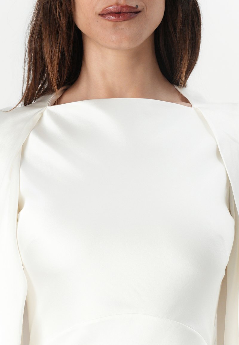 Femme portant une robe écrue à encolure carrée avec des épaules structurées, montrant ses lèvres, son menton, son cou et une partie de ses cheveux.
